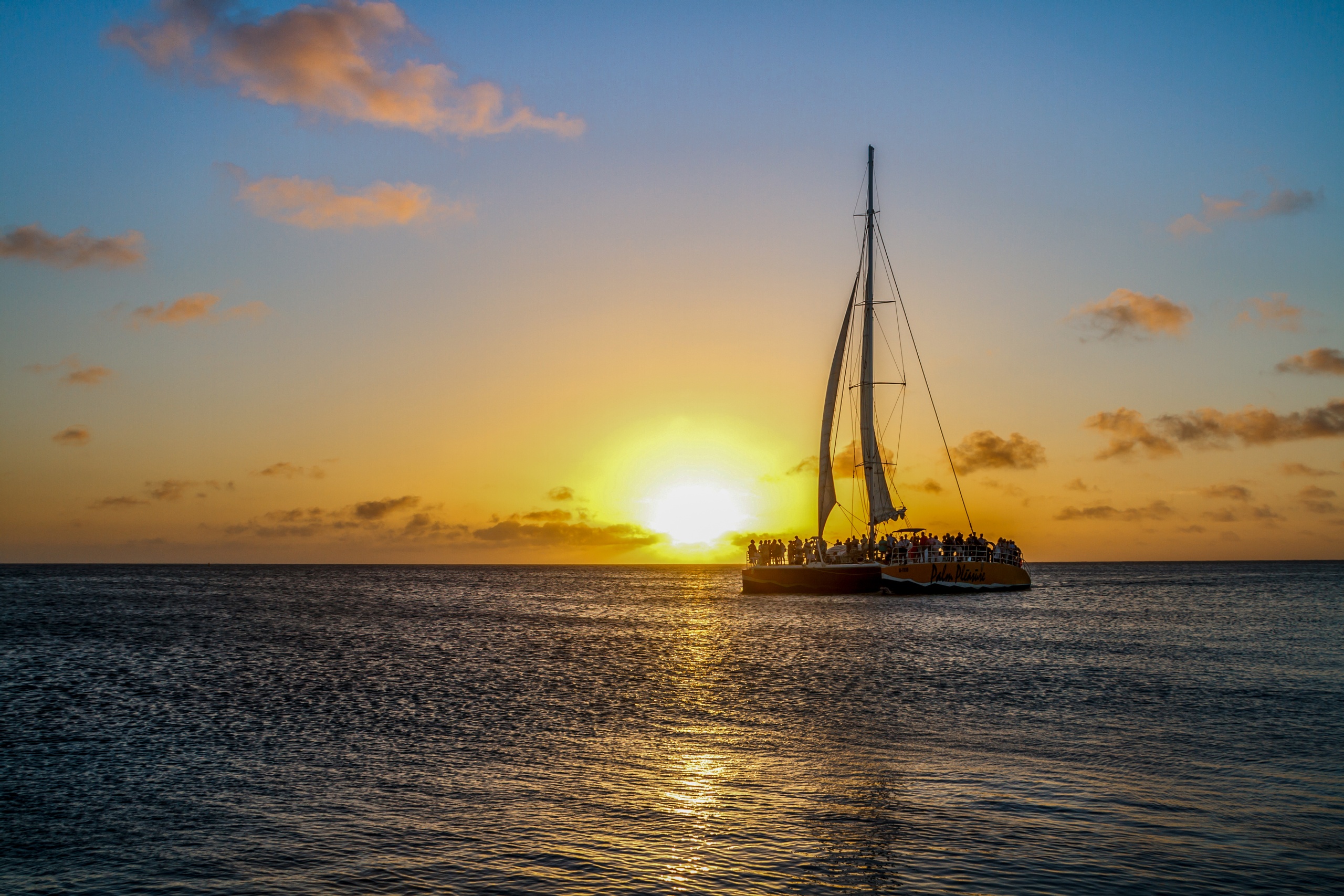 Sailboat on ocean at sunset with a glowing sun and scattered clouds.